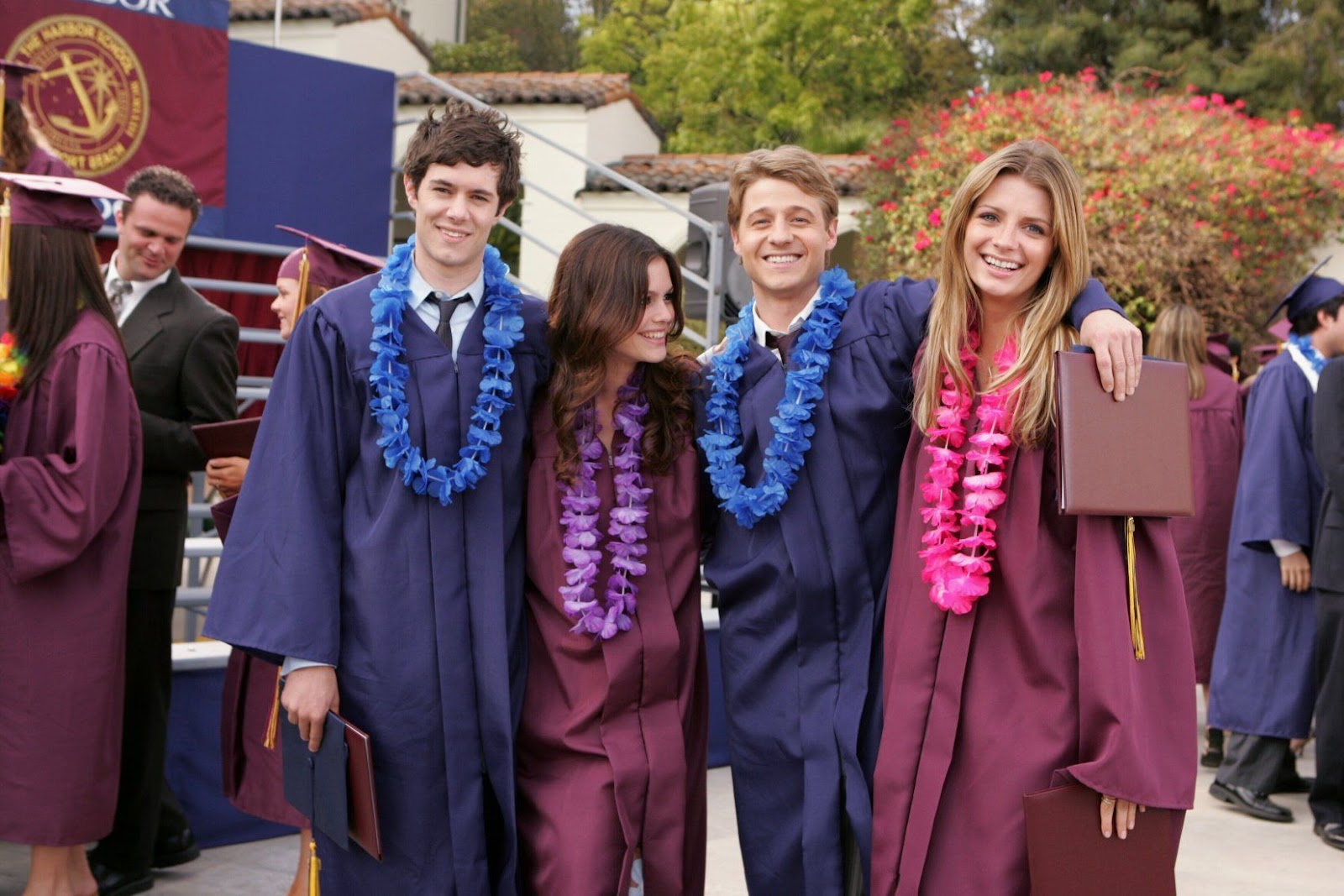 Seth, Ryan, Marissa y Summer posan para la cámara tras su graduación de la Harbor School. Los hombres visten togas azules con collares de flores a juego, mientras que las mujeres llevan togas burdeo con flores rosadas y moradas.
