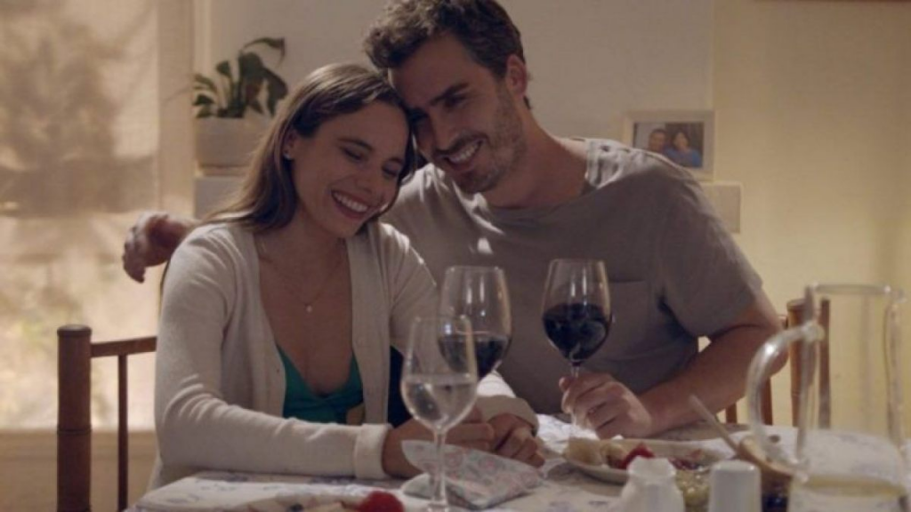 Nicolás Oyarzún y María Gracia Omegna sonrientes y abrazados, compartiendo una copa de vino en una escena íntima y acogedora.